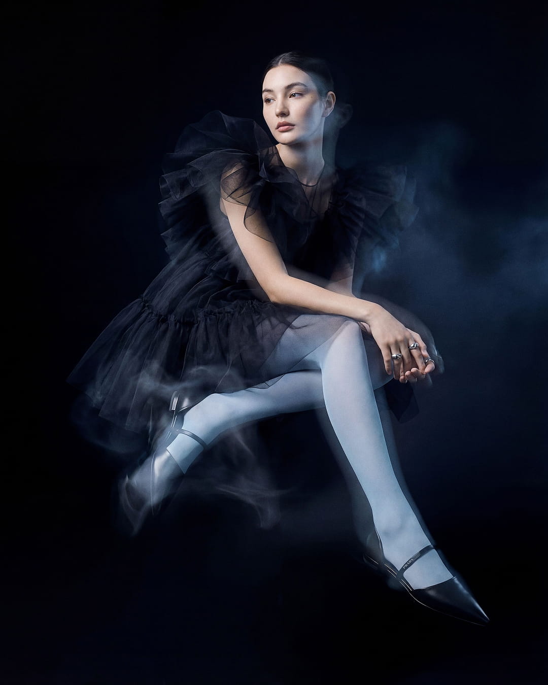 Model in a sheer black tulle dress with white tights and black heels, seated against the dark background with a soft mist effect